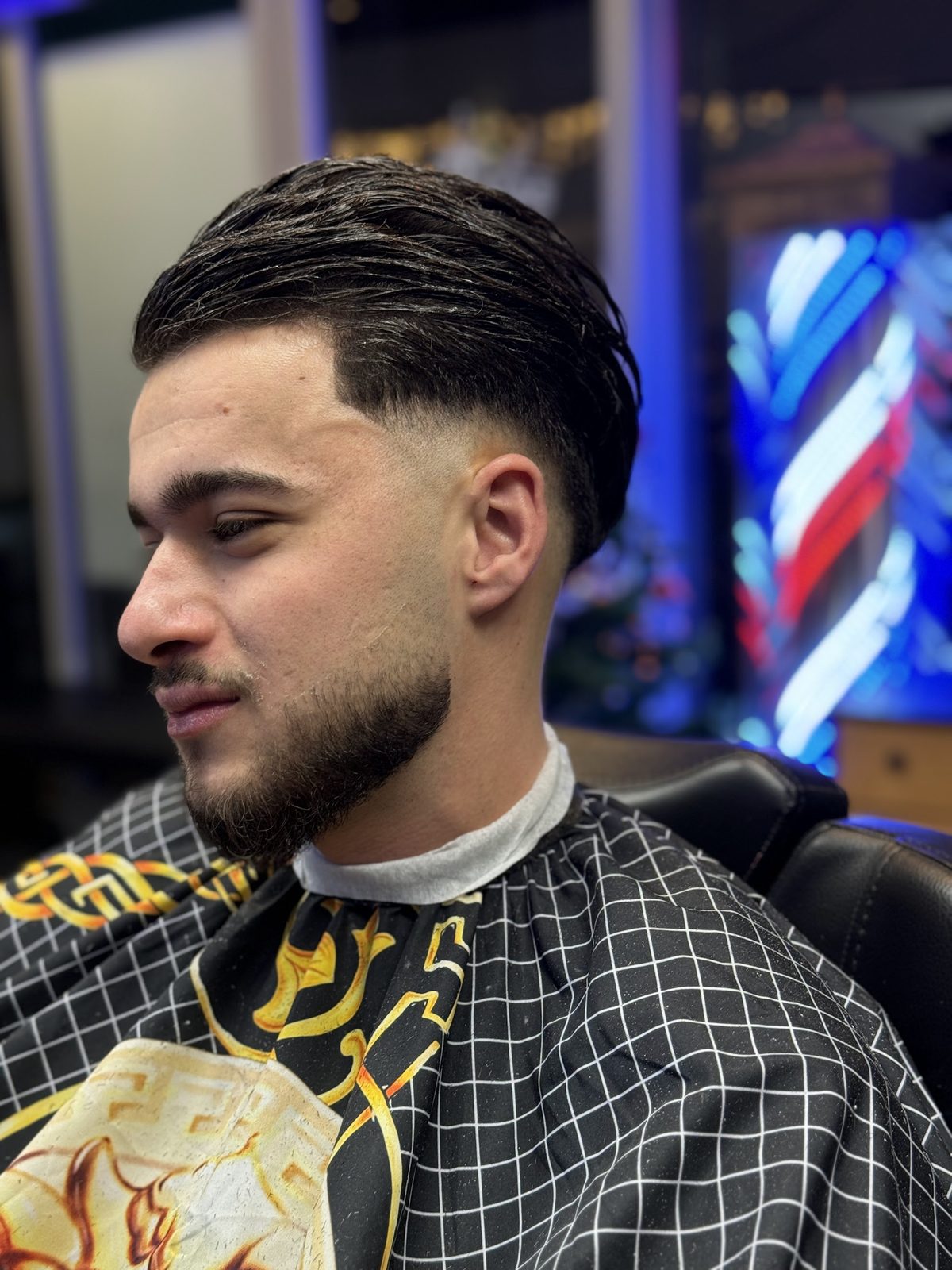 Naar achter gekamd haar met een lage fade en een strak afgelijnde baard, met de barberspaal van de zaak op de achtergrond
